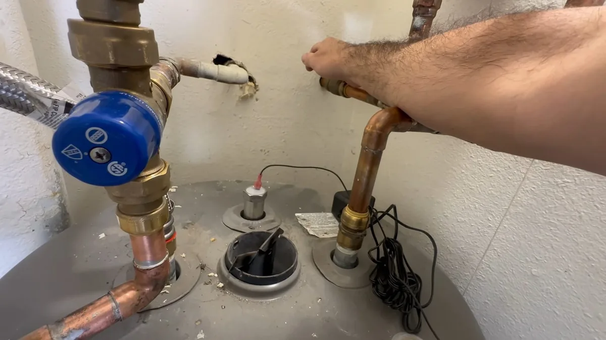 Arm reaching across the top of a gray tank water heater where copper supply lines and a blue valve connect.