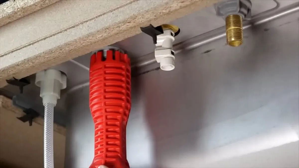 Red basin wrench under a sink basin beside a white supply line and a brass fitting.