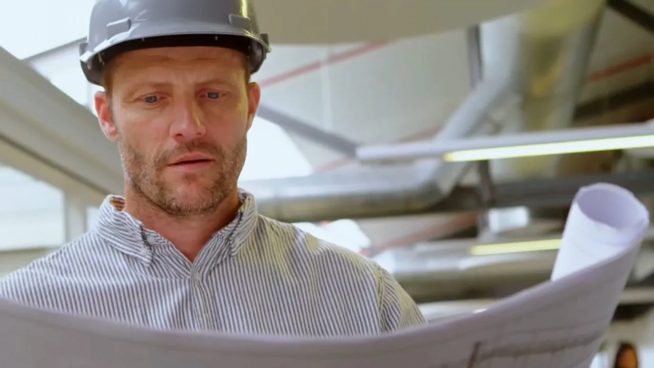 Man in a hard hat looking down at a set of rolled plans, with ducts and pipes overhead.