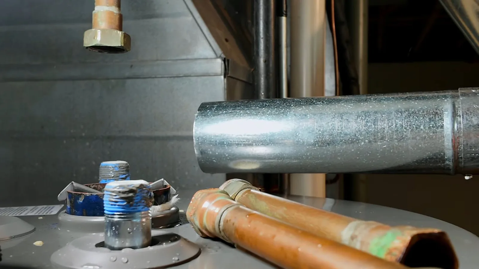 Top of a gas water heater with two copper supply pipes, threaded inlets with blue thread sealant and a steel flue pipe.