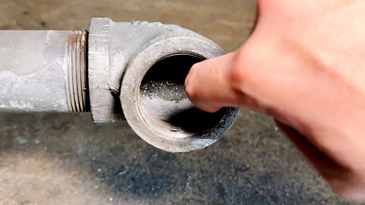 Finger pointing into a galvanized pipe elbow with dark buildup coating the inside.