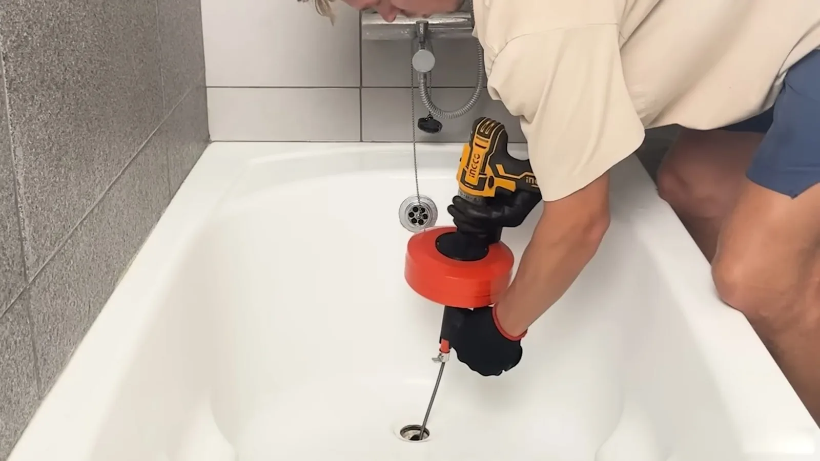 Man leaning over a bathtub with a cordless drill driving a drain auger into the tub drain.