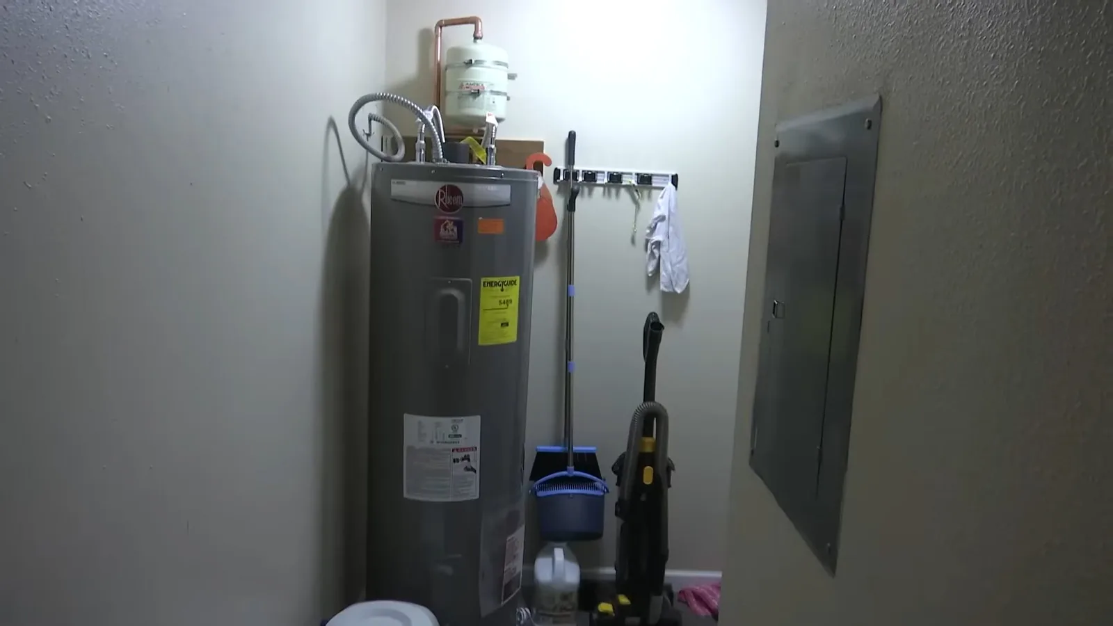 Electric water heater standing in a small utility room beside a gray wall panel, with a broom and dustpan next to it.