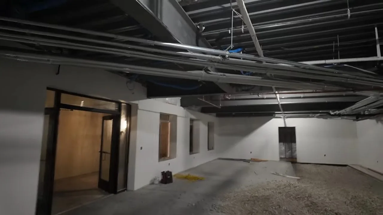 Exposed pipes and ductwork across the ceiling of an unfinished commercial interior with glass doors at the far end.