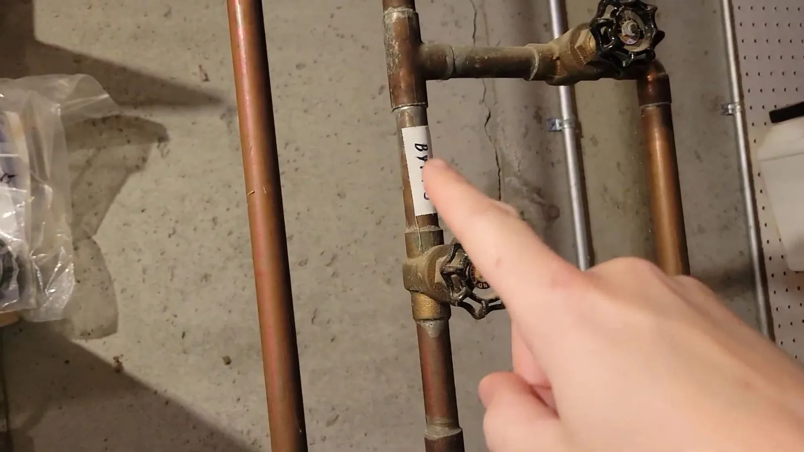 Finger pointing at a gate valve on vertical copper water pipes beside a concrete wall.