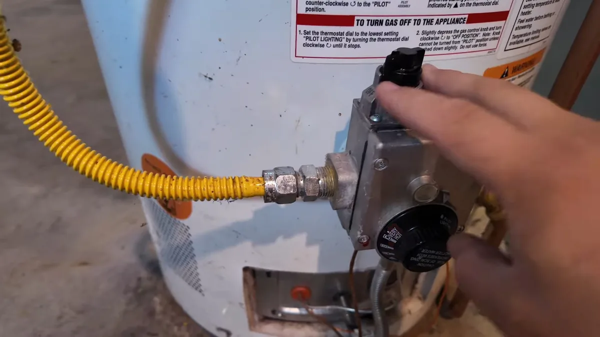 Hand on the gas control valve and temperature dial of a white gas water heater, with a yellow gas line attached.