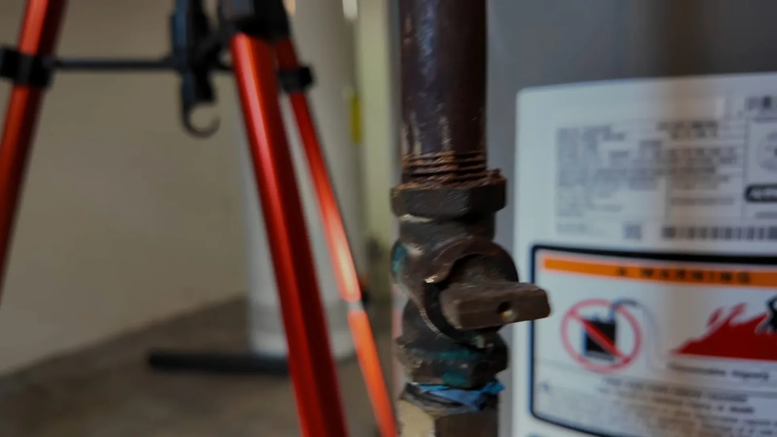 Rusted gas shutoff fitting in front of a water heater with a white warning label, with tripod legs at the left edge.