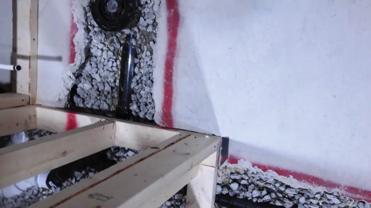 Wooden form on a gravel bed with a black drain pipe and red marker lines against a white wall, before a slab is poured.