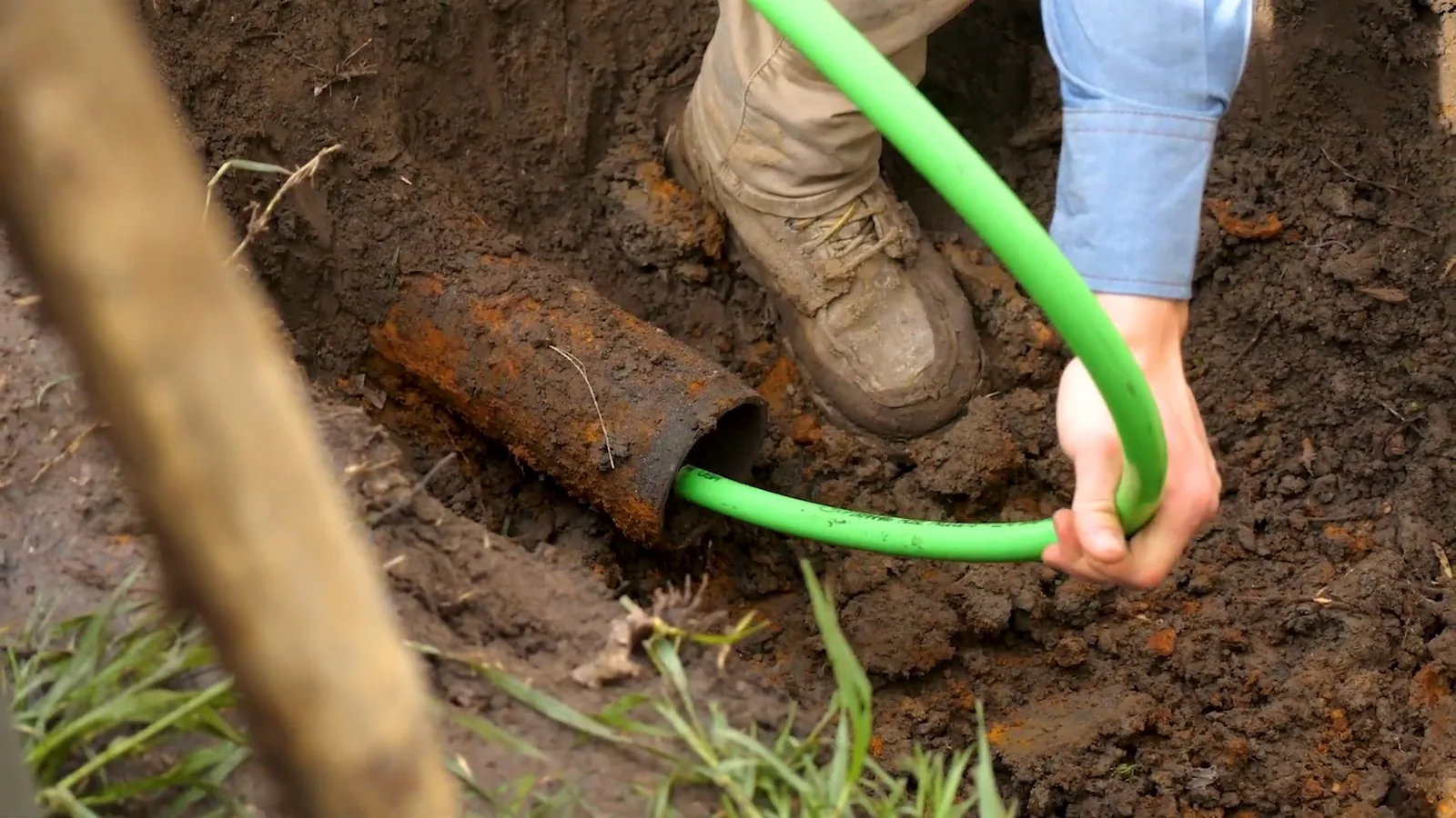 Hand guiding a bright green tube into the end of a rusted pipe in a dug pit, with a boot planted on the soil beside it.