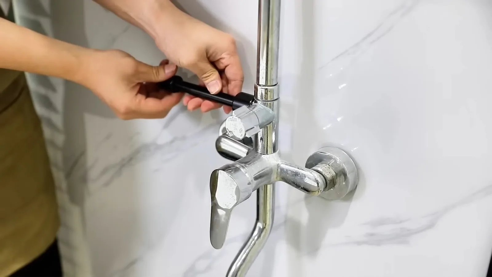 Hands tightening a small fitting on a chrome wall-mounted faucet against white marble-look tile.