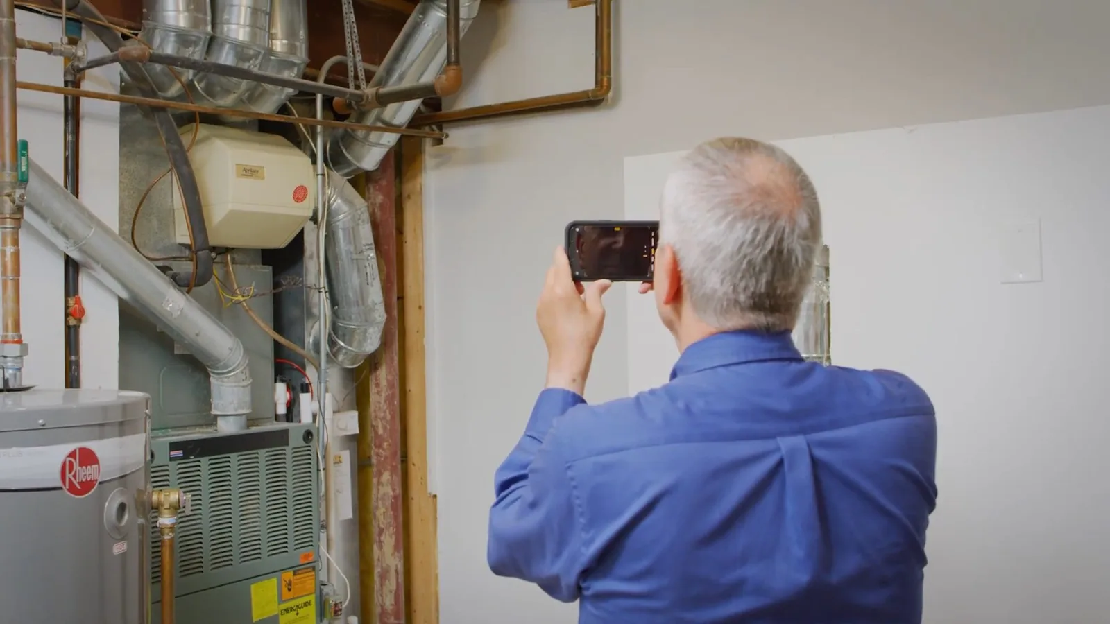Man in a blue shirt holding a phone up to photograph a wall of equipment that includes a water heater and a furnace.