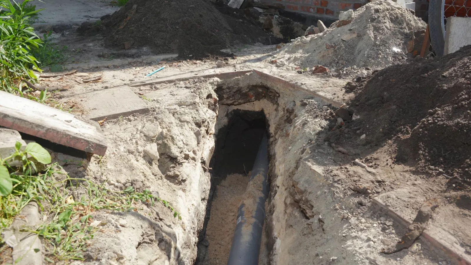 Narrow dirt trench beside a building with a dark pipe in the bottom and soil piled to one side.