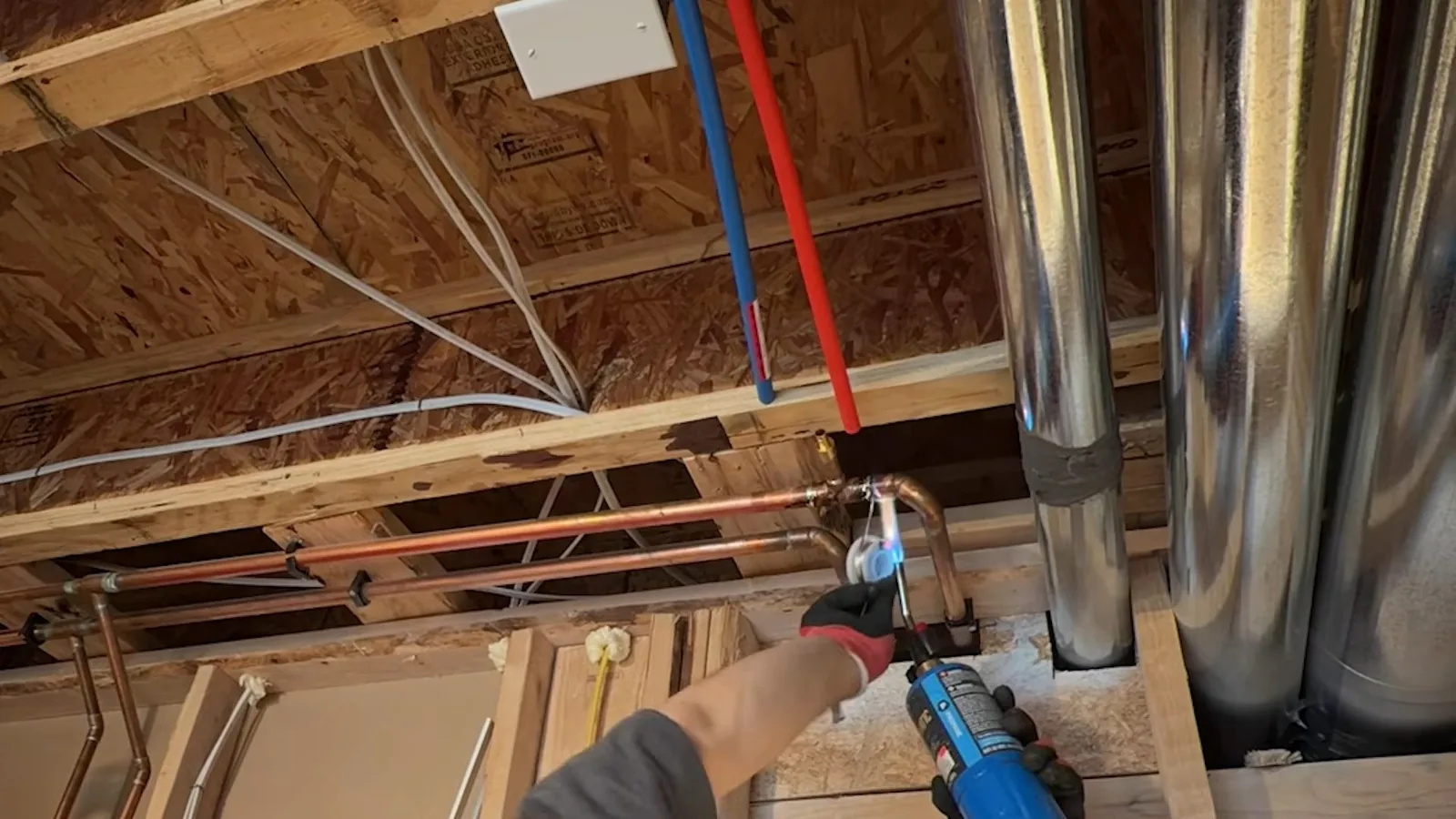Blue and red plastic supply lines and copper pipe running through open basement joists, with a hand holding a torch at a joint.