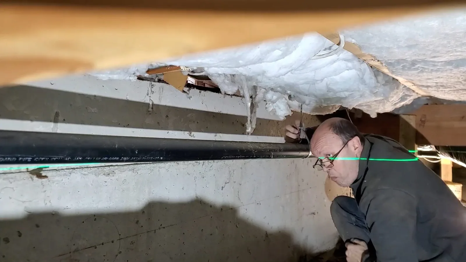 Man crouched in a crawlspace beside a long black pipe under insulated floor joists, a green laser line crossing the pipe.