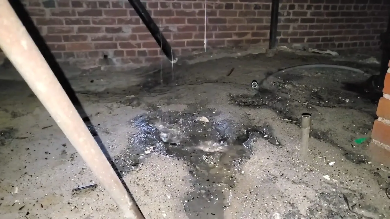 Wet gravel and dark pooled water on a crawlspace floor beneath pipes, with water dripping from above.