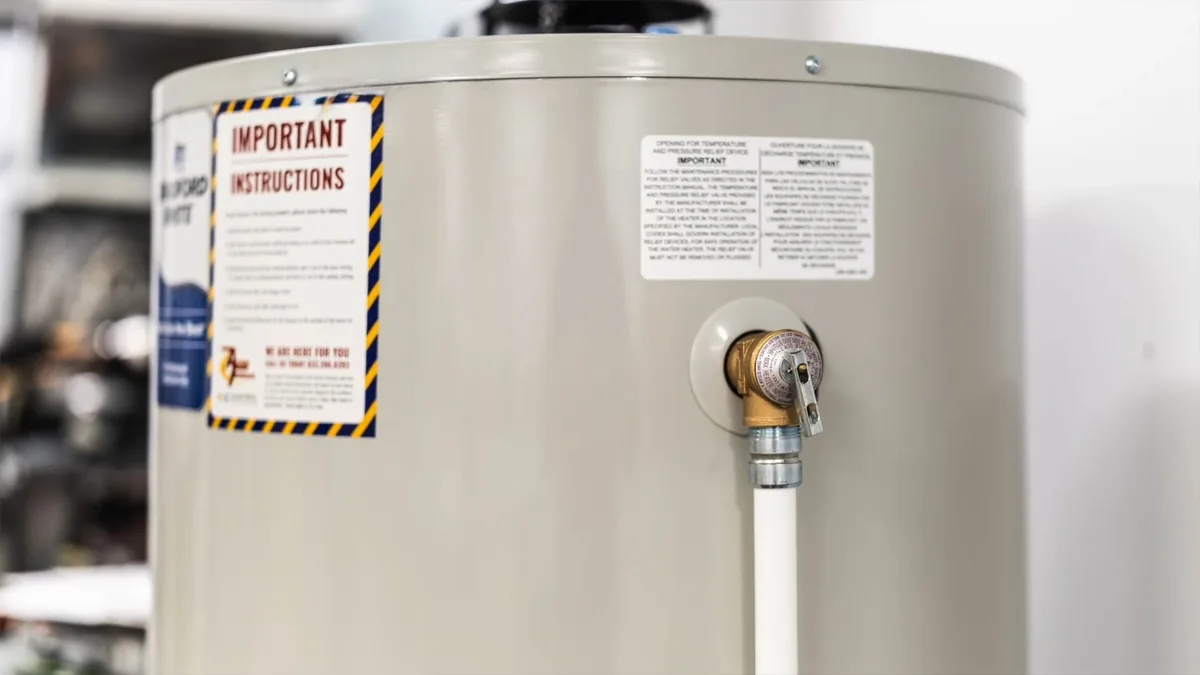 Brass relief valve with a white pipe attached on the side of a cream-colored water heater tank, beside warning labels.