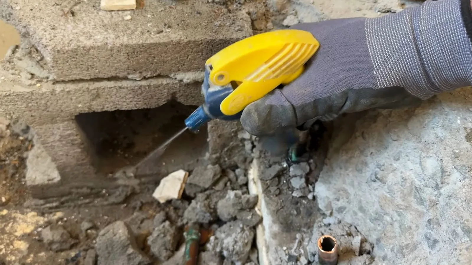 Gloved hand holding a yellow spray head over a hole broken into a concrete slab, with pipe ends in the debris.