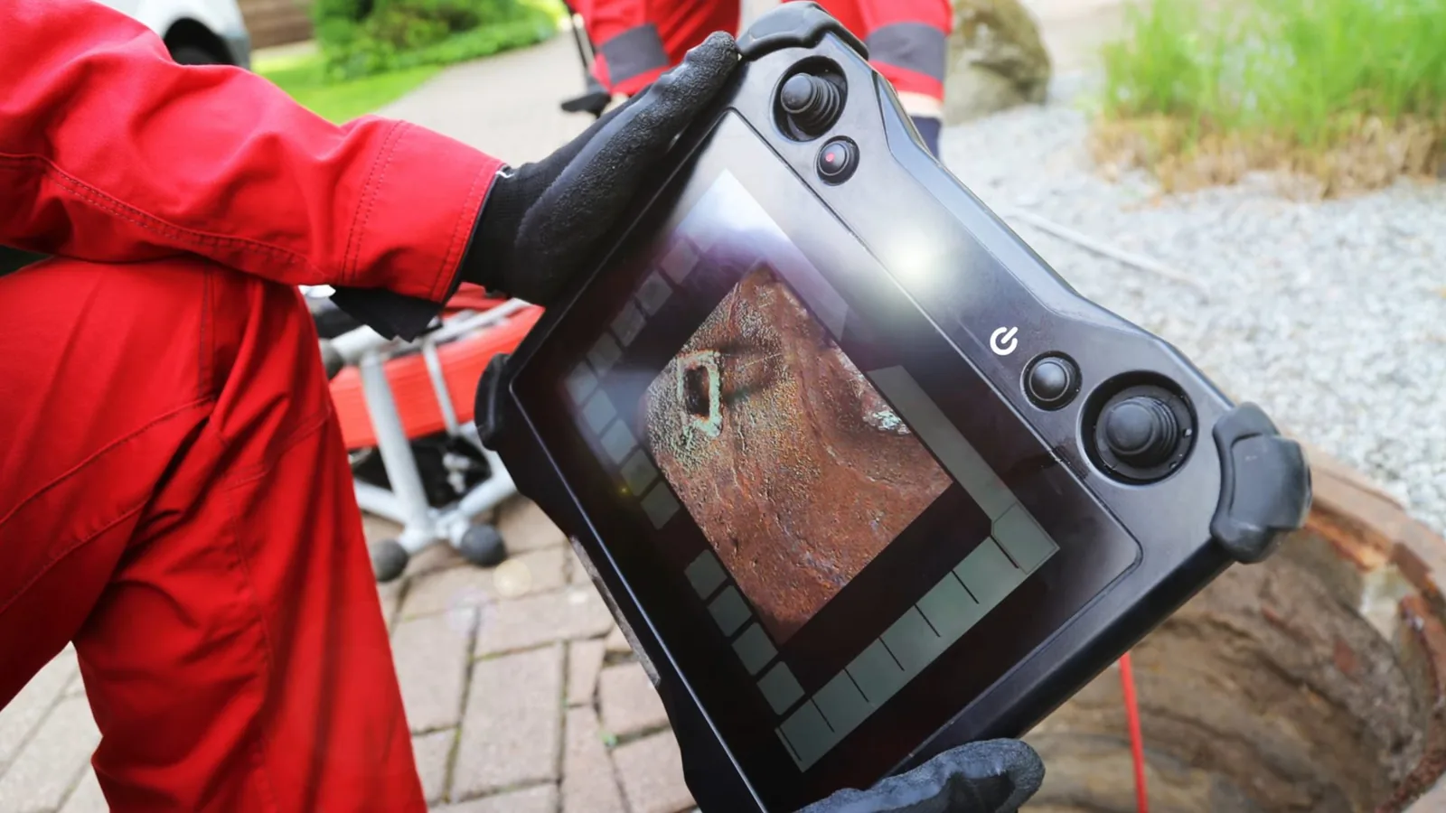 Technician in red coveralls holding a camera monitor that shows the inside of a pipe, beside a ground-level access opening.
