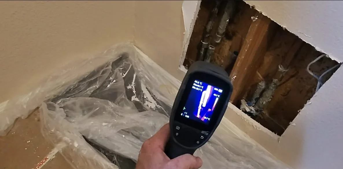 Hand holding a thermal camera at a corner of opened drywall, with pipes visible inside the wall.