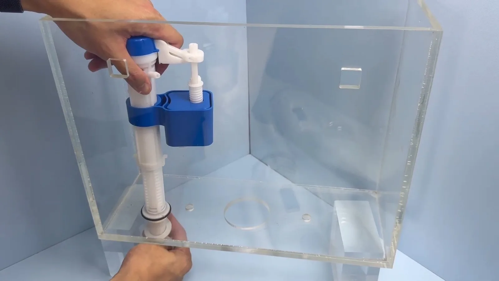 Hands holding a blue and white fill valve assembly inside a clear acrylic toilet tank.
