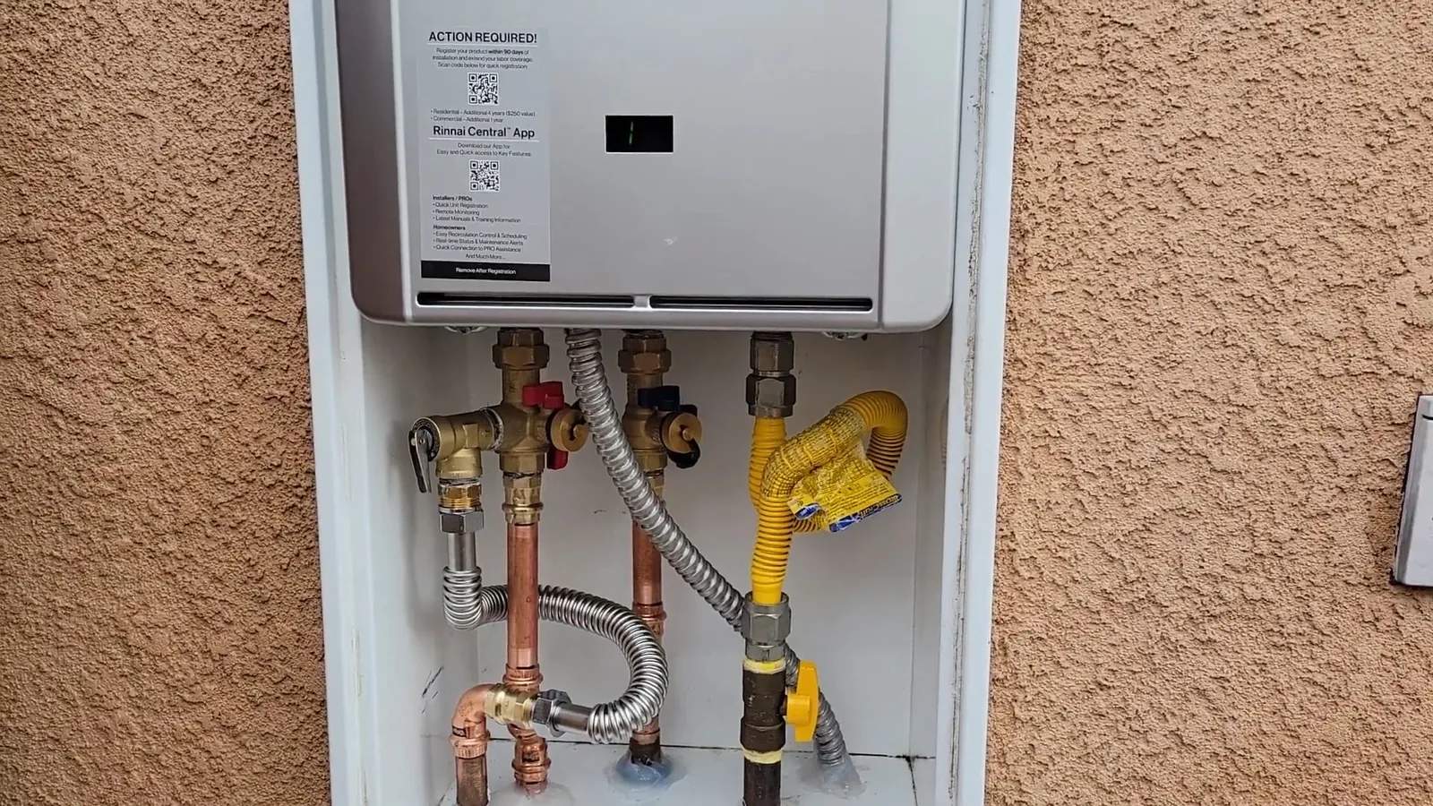 Tankless water heater mounted in a white recess on a stucco wall, with copper pipes, a gas line and valves below it.