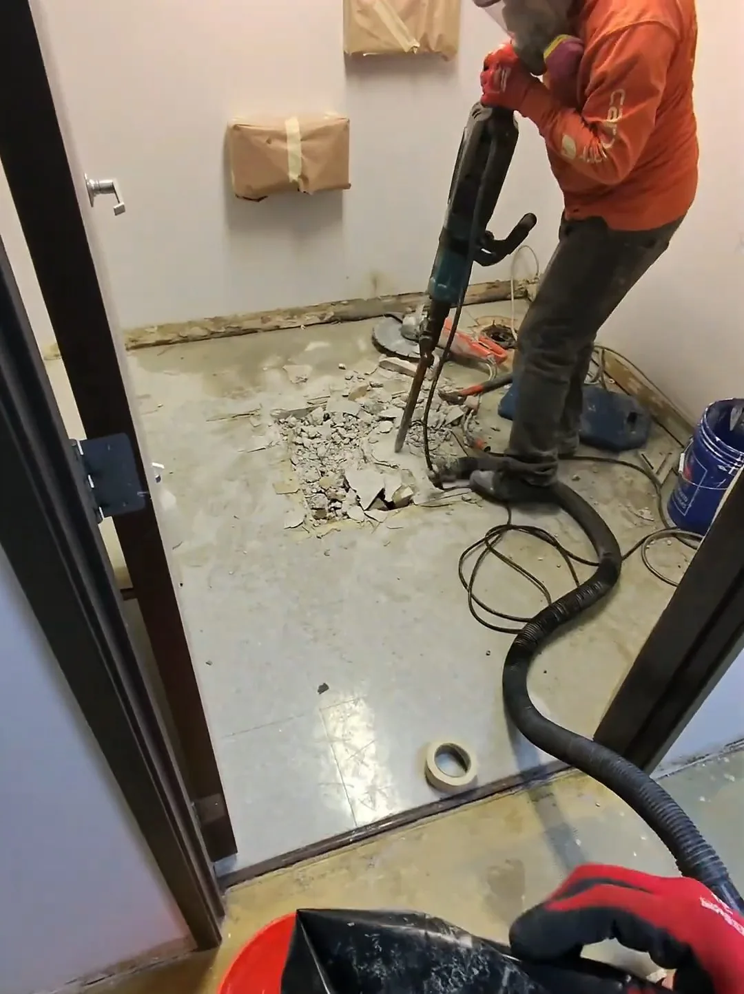 Worker using a breaker hammer on a concrete floor inside a small room, with a vacuum hose beside them.