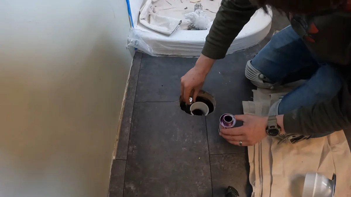 Person kneeling on a bathroom subfloor and fitting a pipe into an open drain flange, with a toilet bowl wrapped in plastic at the upper left.