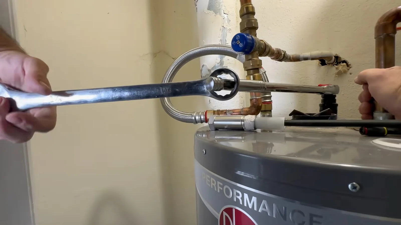 Hands holding a long wrench on a fitting at the top of a gray tank water heater, with a blue valve and braided hose above.